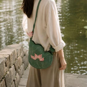 May include: A green shoulder bag shaped like a lily pad with white embroidered veins and a three-dimensional pink lotus flower appliqué on the front. The bag features a matching green twisted strap and a smaller pink lotus bud accent near the top.