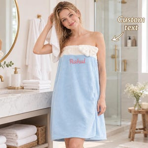 Puede incluir: Una toalla de baño sin tirantes con una sección inferior azul claro y una banda superior color crema con un lazo decorativo. La toalla está bordada con el nombre Rahul en letra cursiva rosa, y una flecha señala el texto con la etiqueta Custom text.