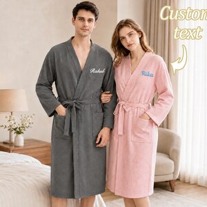 May include: A dark gray waffle-knit robe with the embroidered name Rahul and a light pink waffle-knit robe with the embroidered name Rika. Both robes feature long sleeves, a tie waist, and two front pockets. Text reads Custom text.