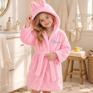 May include: A plush pink hooded bathrobe featuring two small ears on the hood and a tie belt at the waist. The name Olivia is embroidered in blue script on the left chest. The robe is made of a soft, fuzzy material with long sleeves.
