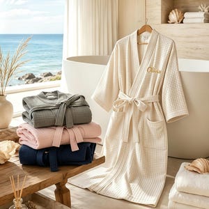May include: A collection of waffle-textured robes in cream, gray, pink, and navy blue. The cream robe hangs on a wooden hanger and features the embroidered name Olivia on the chest. Each robe includes a matching tie belt and two front pockets.