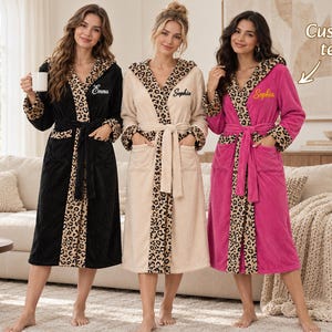 May include: Three plush robes in black, beige, and bright pink, each featuring a leopard print hood, cuffs, and front trim. The robes include a matching tie belt, two front pockets, and personalized embroidery on the chest. Text reads Custom text.
