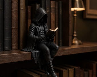 May include: A dark gray figurine of a hooded figure sitting on the edge of a shelf, wearing a long coat and pants while holding an open book. The figure has a shadowed face and is positioned among several rows of vintage-style hardcover books.