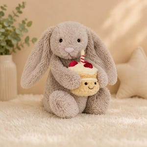 Puede incluir: Un conejo de peluche beige con largas orejas caídas y nariz rosa, sosteniendo un pequeño cupcake de peluche color crema. El cupcake presenta una cereza roja en la parte superior, una vela a rayas y una cara sonriente bordada en la base.