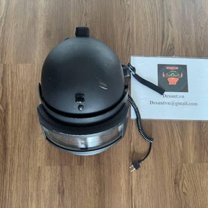 May include: A matte black helmet featuring a clear visor, a top-mounted strap, and a black coiled electrical cord with a two-prong plug. Next to the helmet is a white card with the text Desant.vn and Desantvn@gmail.com printed on it.