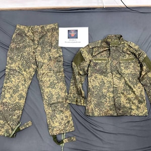 May include: A camouflage uniform set consisting of long trousers and a long-sleeved jacket in shades of green, brown, and black. The jacket features a high collar, chest pockets, and shoulder epaulets. A white card with text is placed between the items.