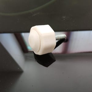 May include: A white, hexagonal-shaped knob with a smooth, rounded top surface. A threaded metal bolt extends from the back of the knob, designed for secure attachment. The component features a clean, matte finish and a simple, functional design.