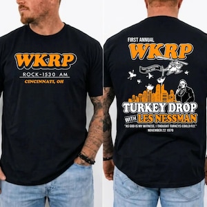 Könnte beinhalten: Ein schwarzes T-Shirt mit dem orange-weißen Schriftzug WKRP ROCK-1530 AM CINCINNATI, OH auf der Vorderseite. Die Rückseite zeigt eine Grafik eines Hubschraubers, einer Skyline und Les Nessman mit dem Text FIRST ANNUAL WKRP TURKEY DROP WITH LES NESSMAN.