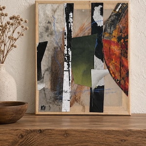May include: A rectangular mixed media collage artwork in a light wood frame featuring layered paper elements in black, white, olive green, and a prominent textured orange arc. The composition includes thin blue lines and the initials KH in black ink.