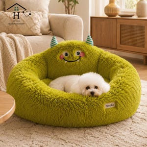 May include: A round, lime green, plush pet bed shaped like a monster with two small striped horns, embroidered eyes, pink cheeks, and a wide smile. A small white dog with curly fur rests inside the bed, which features a white Hevolia brand tag.