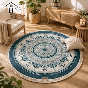 Puede incluir: Una alfombra redonda con un patrón de mandala en color verde azulado y crema, con detalles geométricos intrincados y un borde con flecos. La alfombra tiene una pequeña etiqueta rectangular con el texto Hevolia cosido cerca del borde inferior.