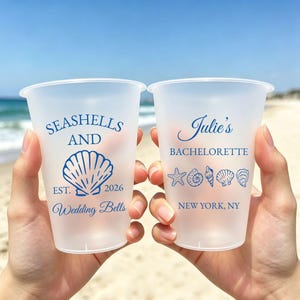 Op de afbeelding: Twee matglazen plastic bekers met blauwe tekst en afbeeldingen. De linkerbeker toont SEASHELLS AND EST. 2026 Wedding Bells met een schelppictogram. De rechterbeker toont Julie's BACHELORETTE NEW YORK, NY met een rij van zeester- en schelppictogrammen.