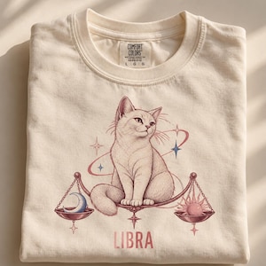 Könnte beinhalten: Ein cremefarbenes T-Shirt mit einer Grafik einer Katze, die auf einer Waage sitzt. Die Waage trägt einen blauen Halbmond auf der einen und eine rosa Sonne auf der anderen Seite. Das Wort LIBRA ist in Rosa unter der Waage gedruckt. Comfort Colors Etikett.