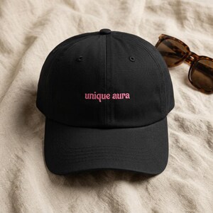 May include: A black baseball cap featuring the embroidered text unique aura in pink thread on the front panel, accompanied by a pair of brown tortoiseshell sunglasses with dark lenses positioned to the right of the cap.
