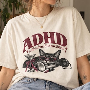 May include: An off-white t-shirt featuring a graphic of a cat wearing red sunglasses, resting on a book next to a martini glass with a red bow, a lipstick, and headphones. The text reads ADHD a diva has distractions in dark red lettering.