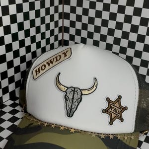 Puede incluir: Una gorra de camionero con panel frontal blanco, visera de camuflaje verde oliva y parte trasera de malla. El frente presenta tres parches: una etiqueta beige 'HOWDY', un cráneo de toro central y una estrella de sheriff dorada con el texto 'YALL'.