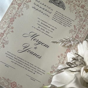 Pode incluir: Um certificado de casamento na cor creme com uma delicada borda floral em tons de rosa e marrom. O documento inclui caligrafia árabe, texto em inglês detalhando a união de Meryem e Younes, e linhas para as assinaturas.