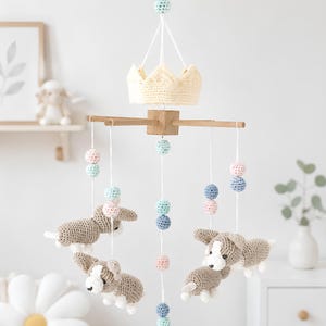 May include: A hanging mobile featuring a central wooden crossbar with a cream-colored crocheted crown on top. Four crocheted dog figures in shades of tan and white hang from white strings, interspersed with small pastel blue, pink, and white yarn balls.