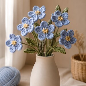 Puede incluir: Un ramo de seis flores de ganchillo con pétalos azul claro y centros amarillos, que incluye hojas y tallos verdes tejidos. Las flores están dispuestas en un jarrón de cerámica de color blanco roto, con acabado mate y liso, cuello estrecho.