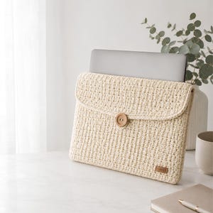 Puede incluir: Una funda de ganchillo para portátil en color crema con un patrón de rejilla texturizado, una solapa plegable asegurada con un botón de madera redondo y una pequeña etiqueta rectangular de cuero marrón en la esquina inferior derecha.