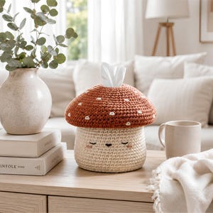 May include: A crochet tissue box cover shaped like a mushroom with a rust-colored cap featuring small white embroidered spots and a cream-colored base with a stitched face showing closed eyes, a small mouth, and pink cheeks, with a white tissue.