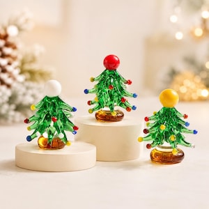 Pode incluir: Três árvores de Natal em miniatura de vidro com ramos verdes e ornamentos de contas de vidro coloridas. Cada árvore tem uma base de vidro maciço e uma grande esfera no topo, branca, vermelha e amarela, sobre pedestais circulares.