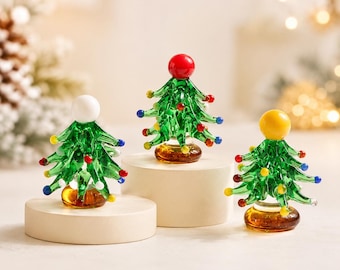 Könnte beinhalten: Drei Miniatur-Weihnachtsbäume aus Glas mit grünen Zweigen und bunten Glasperlenverzierungen. Jeder Baum hat einen massiven Glassockel und eine große Kugel an der Spitze in Weiß, Rot und Gelb, die auf runden Podesten platziert sind.