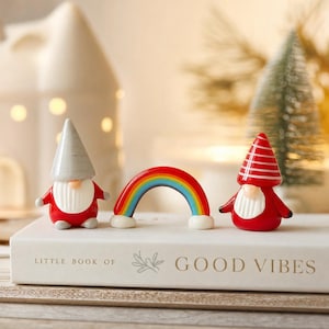 May include: Two ceramic gnome figurines and a small rainbow arch sit on a white book titled Little Book of Good Vibes. One gnome wears a grey pointed hat, the other a red and white striped hat. Both have red bodies and white beards.