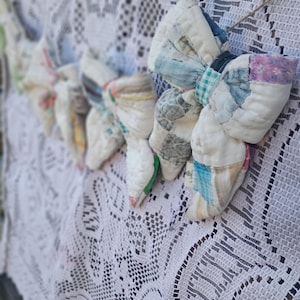 May include: A garland of multiple stuffed fabric bows made from quilted patchwork scraps in various colors and patterns, including white, blue, and pink, suspended on a thin string against a white crocheted lace fabric background.
