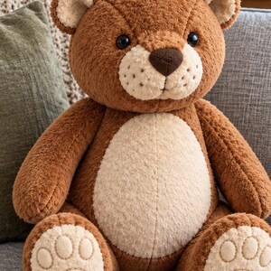 Puede incluir: Un oso de peluche con pelaje marrón y vientre, hocico e interior de las orejas de color crema. El oso presenta ojos oscuros y redondos, una nariz cosida de color marrón oscuro y almohadillas color crema con detalles circulares cosidos.