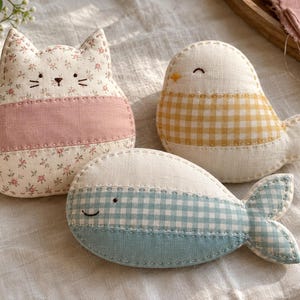 May include: Three stuffed fabric ornaments shaped like a cat, a bird, and a whale. The cat features a floral print with a pink band, the bird has a yellow gingham pattern, and the whale displays a light blue gingham design with stitched details.