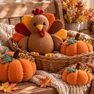 Peut inclure: Une peluche en forme de dinde avec un corps marron, un bec jaune, une caroncule rouge et des plumes de queue en feutrine multicolores, accompagnée de trois citrouilles en tissu orange avec des feuilles et des tiges en feutrine verte.