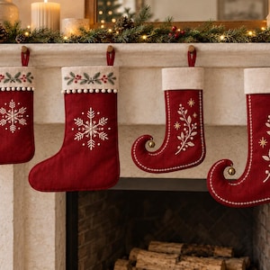 Könnte beinhalten: Vier tiefrote Weihnachtsstrümpfe aus Stoff hängen in einer Reihe. Zwei Strümpfe zeigen weiße gestickte Schneeflocken, während die anderen beiden geringelte Spitzen mit goldenen Glöckchen und weißen Zweigen haben.