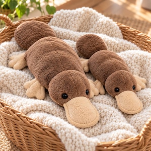 Puede incluir: Dos peluches de ornitorrinco con suave pelaje marrón, picos color crema y patas palmeadas. Los juguetes tienen ojos negros redondos y colas planas, descansando sobre una tela blanca texturizada dentro de una cesta de mimbre.