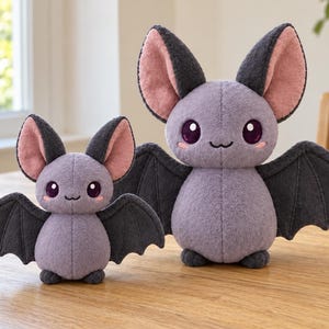 May include: Two plush bat toys in different sizes, both featuring light purple bodies, dark gray wings, and large ears with pink inner lining. Each bat has dark purple eyes, a small stitched mouth, and pink blush marks on its cheeks.