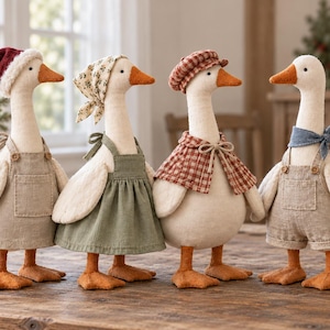 Op de afbeelding: Vier pluche ganzenfiguren staan op een rij. De eerste draagt een rode kerstmuts en een beige tuinbroek. De tweede draagt een hoofddoek met bloemenprint en een saliegroene schortjurk. De derde draagt een rood geruite pet en een bijpassende cape.