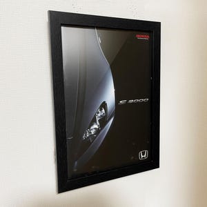 May include: A rectangular black frame containing a poster of a dark grey Honda S2000 car. The poster features the Honda logo, the text Honda The Power of Dreams, and the model name S2000 in white lettering against a black background.