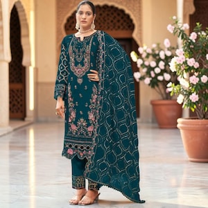 May include: A teal long-sleeved tunic with intricate floral embroidery in pink and gold, paired with matching straight-leg trousers and a coordinating teal dupatta featuring a gold geometric pattern, worn with a pearl necklace and sandals.