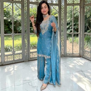 May include: A light blue three-piece ensemble consisting of a long-sleeved tunic with intricate silver embroidery around the neckline, matching wide-leg trousers, and a coordinating sheer dupatta draped over one arm, all featuring a subtle metallic sheen.