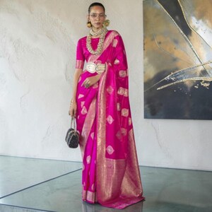 May include: A vibrant fuchsia saree featuring a repeating gold diamond pattern and a wide, ornate gold border. The ensemble includes a matching fuchsia blouse, a layered gold beaded necklace, a gold waist belt, and a small black structured handbag.