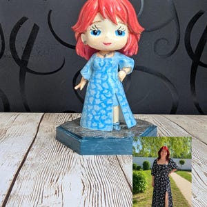 Könnte beinhalten: Eine individuelle Figur mit leuchtend rotem Haar und blauen Augen, die ein hellblaues Kleid mit weißem Blumenmuster trägt. Die Figur steht auf einem dunkelblauen sechseckigen Sockel und wird neben einem Foto einer Person präsentiert.