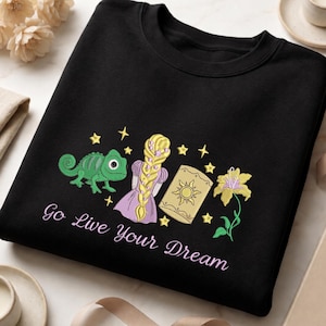 May include: A black crewneck sweatshirt featuring an embroidered design of a green chameleon, a figure with a long blonde braid, a yellow lantern with a sun emblem, a yellow flower, and small stars, with the text Go Live Your Dream below.