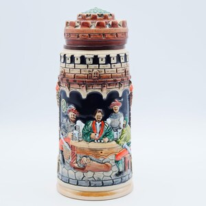 May include: A ceramic beer stein featuring a relief design of four figures seated at a table within a castle setting. The lid is shaped like a brown stone turret. The scene includes a dog resting under the table, with painted details in brown, green, red, and cream tones against a dark background.