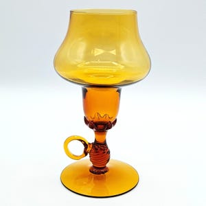 May include: An amber-colored glass candle holder featuring a wide, bowl-shaped top, a central stem with a decorative scalloped collar, a small circular side handle, and a sturdy, flat circular base. The glass is translucent and smooth.