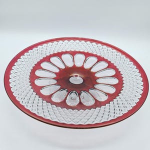 May include: A round glass serving plate featuring a deep red border and a central section with twelve clear, petal-shaped indentations. The outer rim displays a textured diamond-cut pattern, while the center remains smooth and transparent.