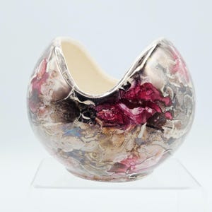 May include: A rounded ceramic vessel with a deep, U-shaped cutout at the top. The exterior features a marbled, abstract pattern in shades of deep magenta, metallic bronze, and gray, while the interior is a smooth, solid off-white color.