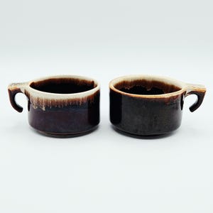 May include: Two ceramic mugs featuring a deep brown glaze with a contrasting cream-colored drip pattern along the upper rim. Each mug has a wide, cylindrical body and a sturdy, angular handle extending from the side of the vessel.