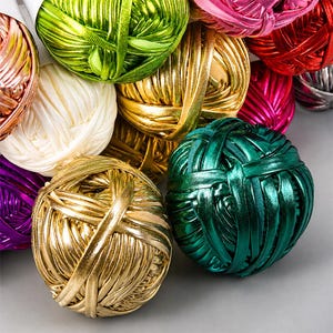 May include: A collection of round yarn balls featuring a metallic, reflective finish in various vibrant colours including gold, emerald green, lime green, white, red, magenta, and purple. Each ball is wound with a smooth, ribbon-like synthetic fibre.