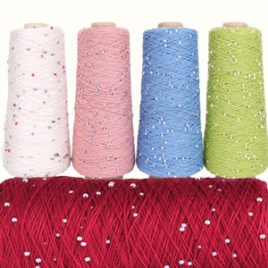 May include: Four upright cones of yarn in white, pink, blue, and lime green, each speckled with tiny colourful beads, positioned behind a large horizontal spool of deep red yarn also featuring small white beads throughout the thread.