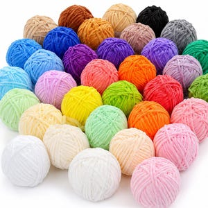May include: A collection of numerous spherical balls of soft, plush yarn arranged in a dense cluster. The yarn balls feature a wide variety of solid colors, including white, cream, yellow, pink, red, orange, green, blue, purple, brown, gray, and black.